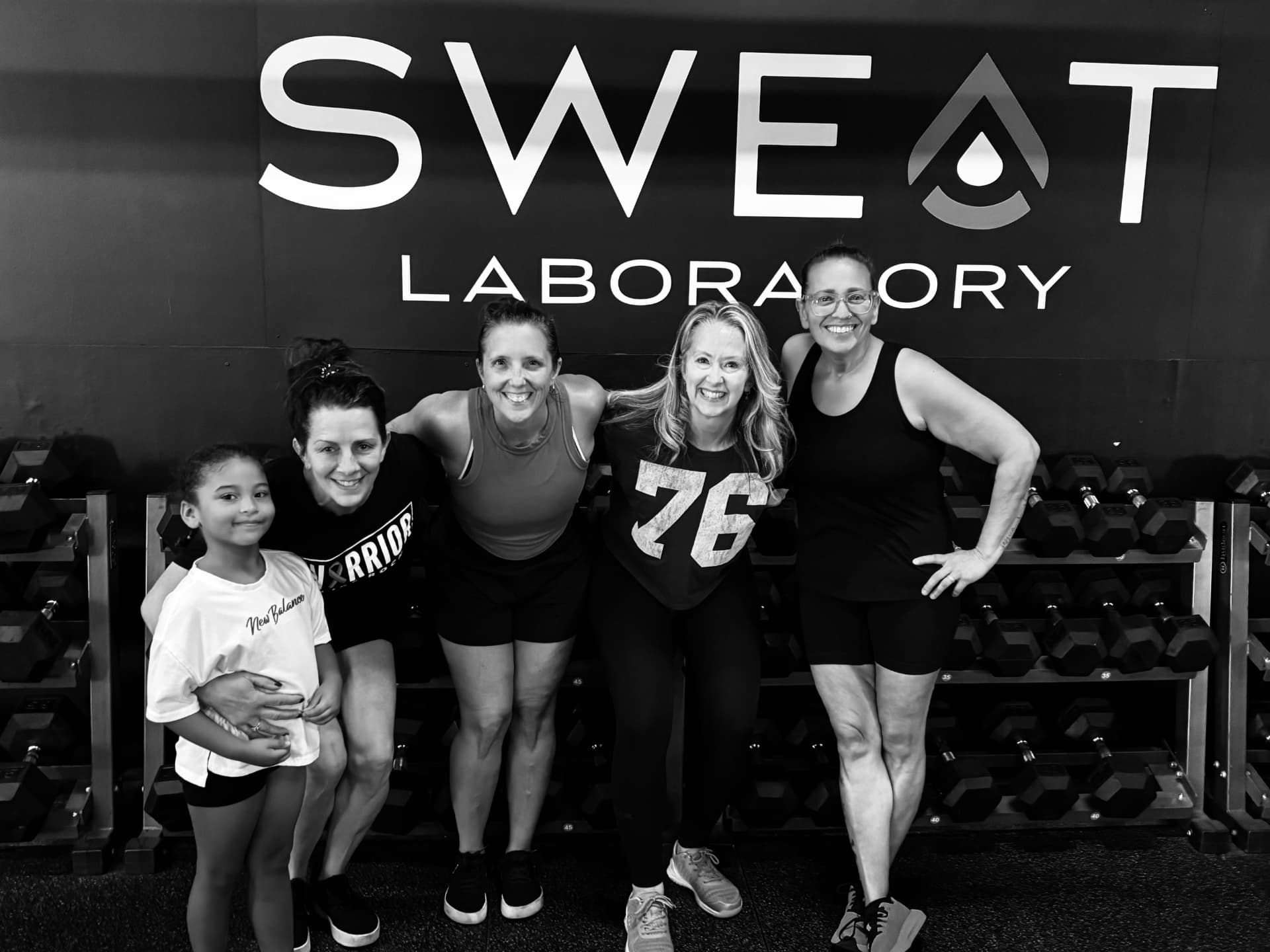 Members and a child smiling together in front of the SWEAT wall